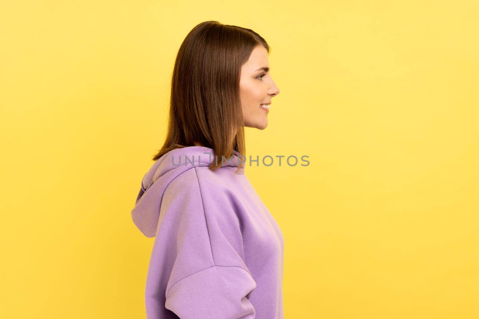 Side view of happy successful woman standing, looking ahead with charming and joyous toothy smile, expressing happiness, wearing purple hoodie. Indoor studio shot isolated on yellow background.