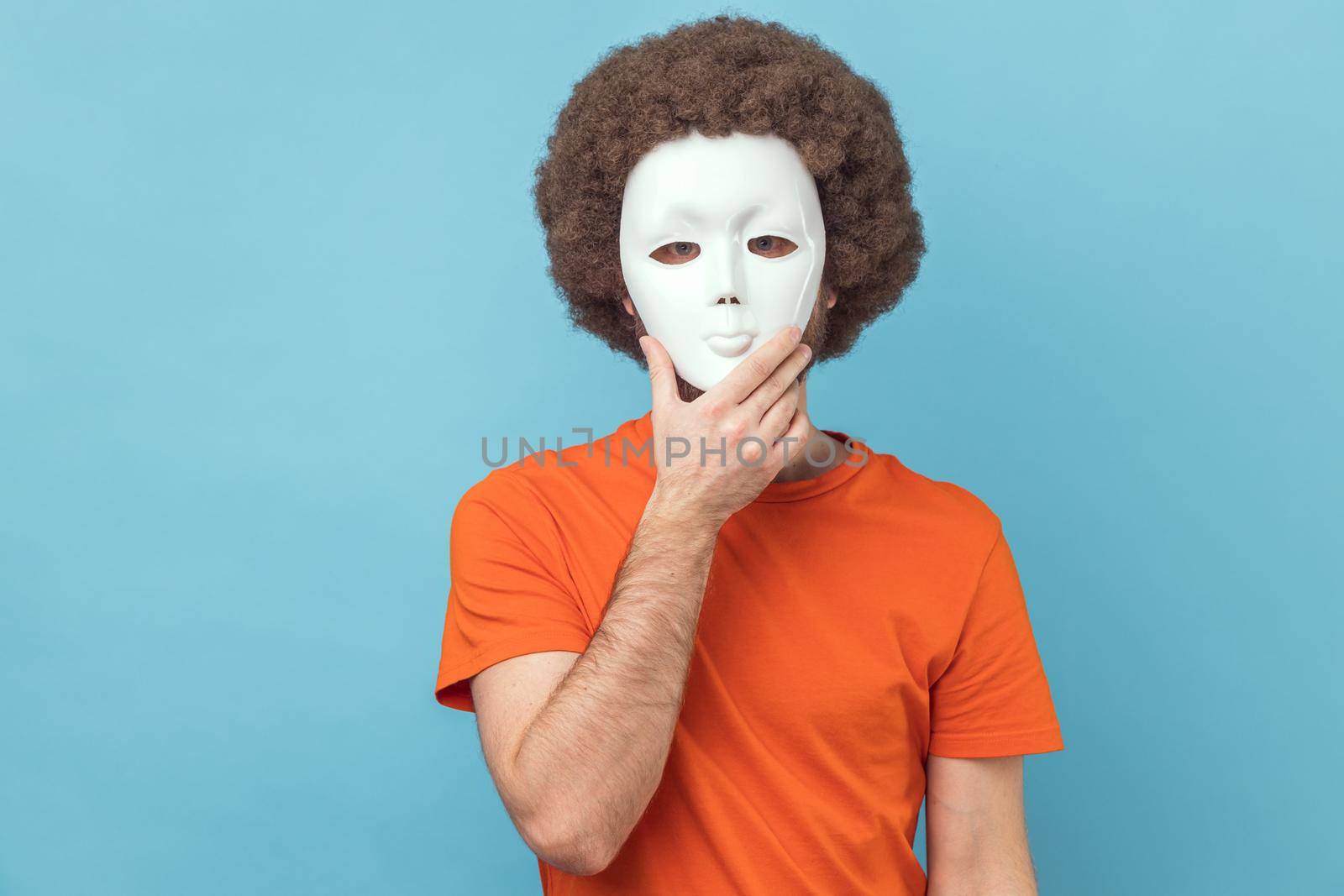 Portrait of man with Afro hairstyle wearing orange T-shirt covering her face with white mask, hiding her real personality, anonymity. Indoor studio shot isolated on blue background.