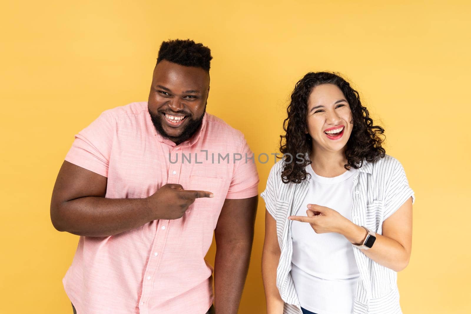 Portrait of funny positive young couple in casual clothing standing together, pointing fingers at each other and looking at camera, laughing. Indoor studio shot isolated on yellow background.