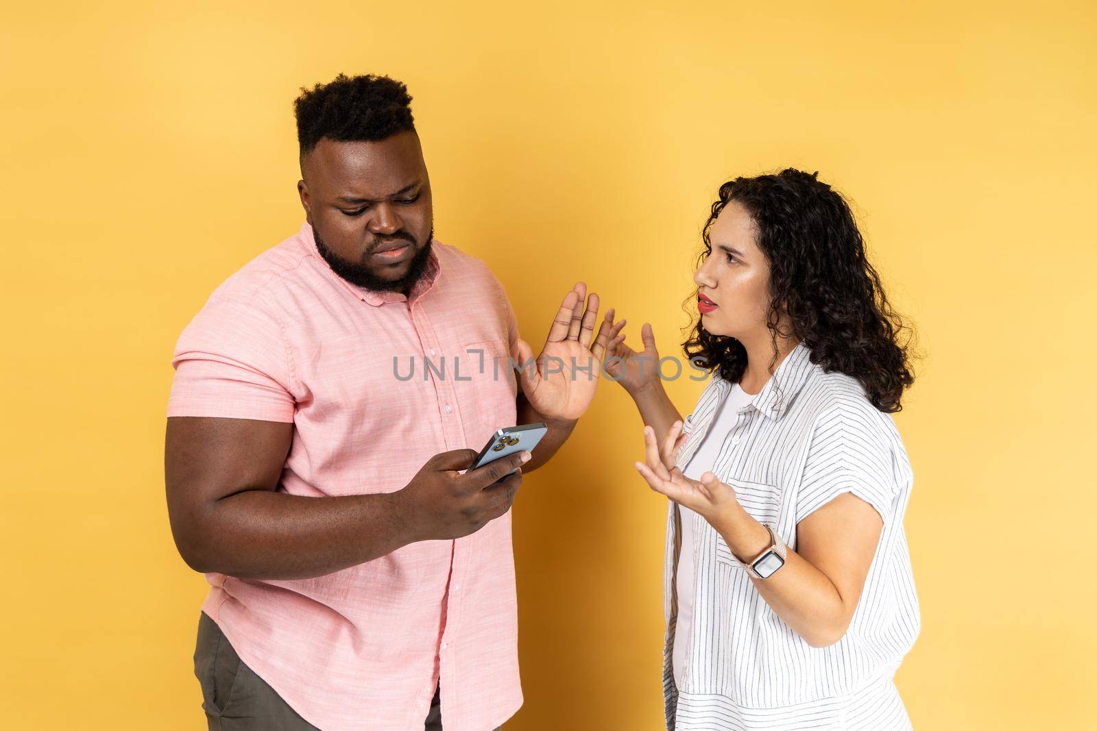 Young couple in casual clothing standing together, man using cell phone and ignoring her wife, woman trying to attract his attention and screaming. Indoor studio shot isolated on yellow background.