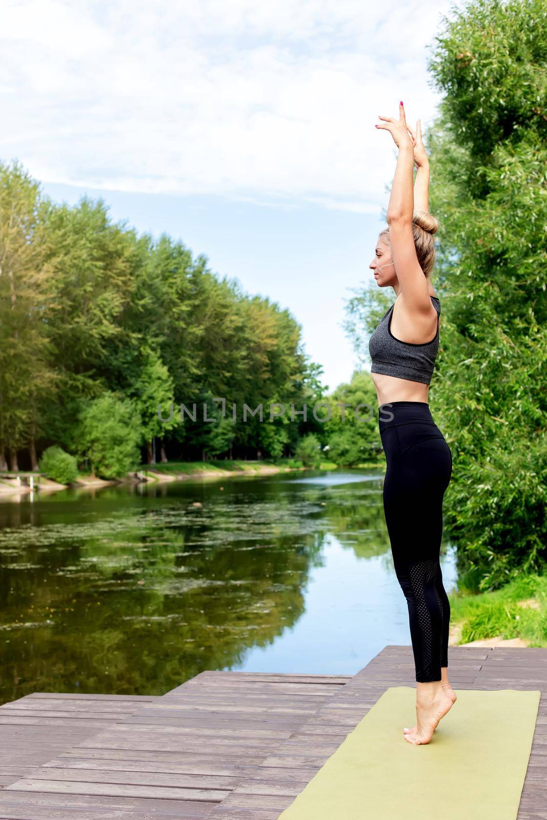 A slender woman stands on a green rug, on a wooden platform by a pond in the park, in summer, with her hands up, doing yoga. Copy space. Vertical