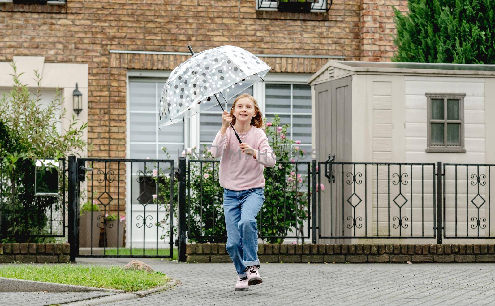 Preteen girl running with umbrella outdoors. Pretty child kid portrait in rainy day at street