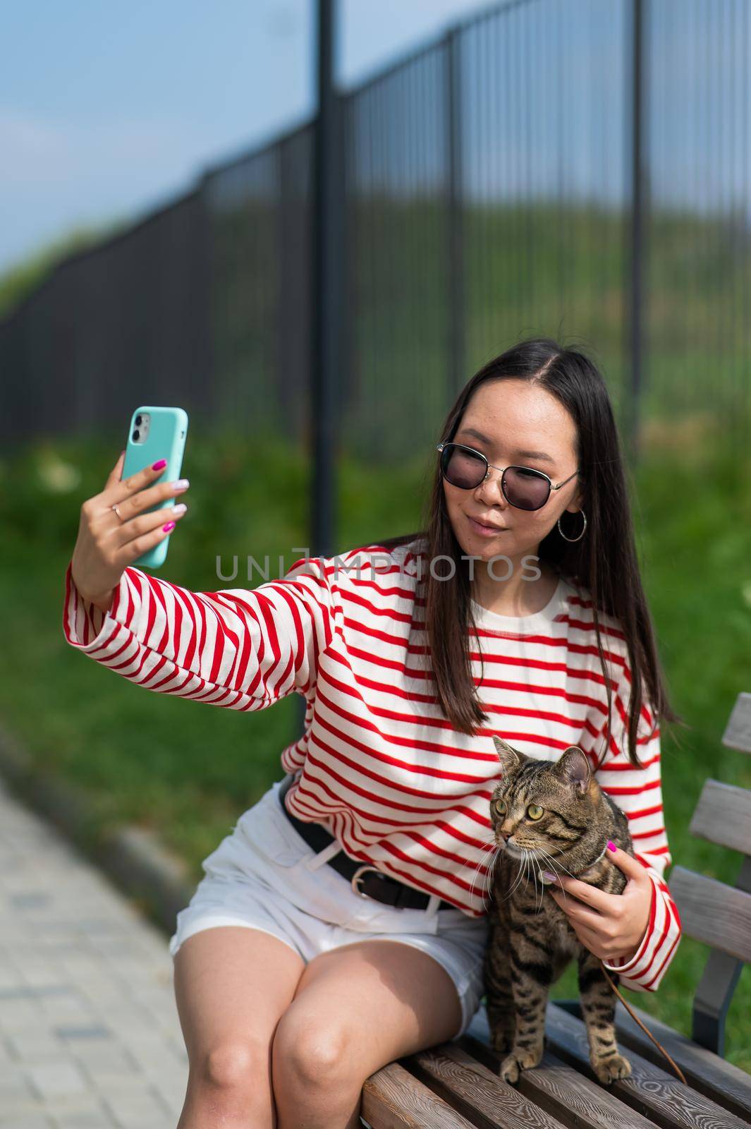 Young woman sits on a bench with a tabby cat and takes a selfie on a smartphone outdoors. by mrwed54