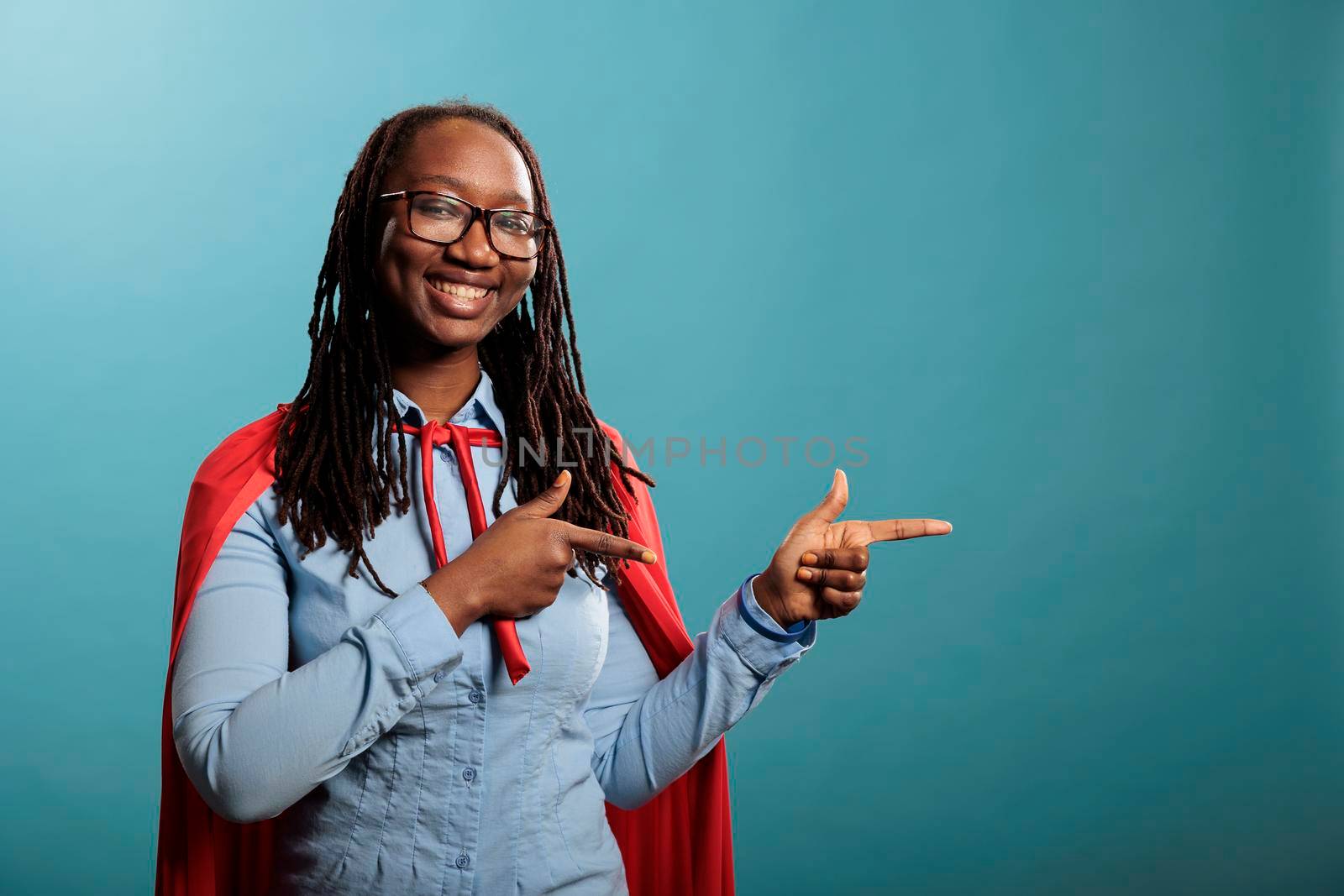 Portrait of young adult african american superhero woman poiting fingers to right while smiling at camera. Beautiful and confident looking person wearing mighty hero cape showing ad placement space.