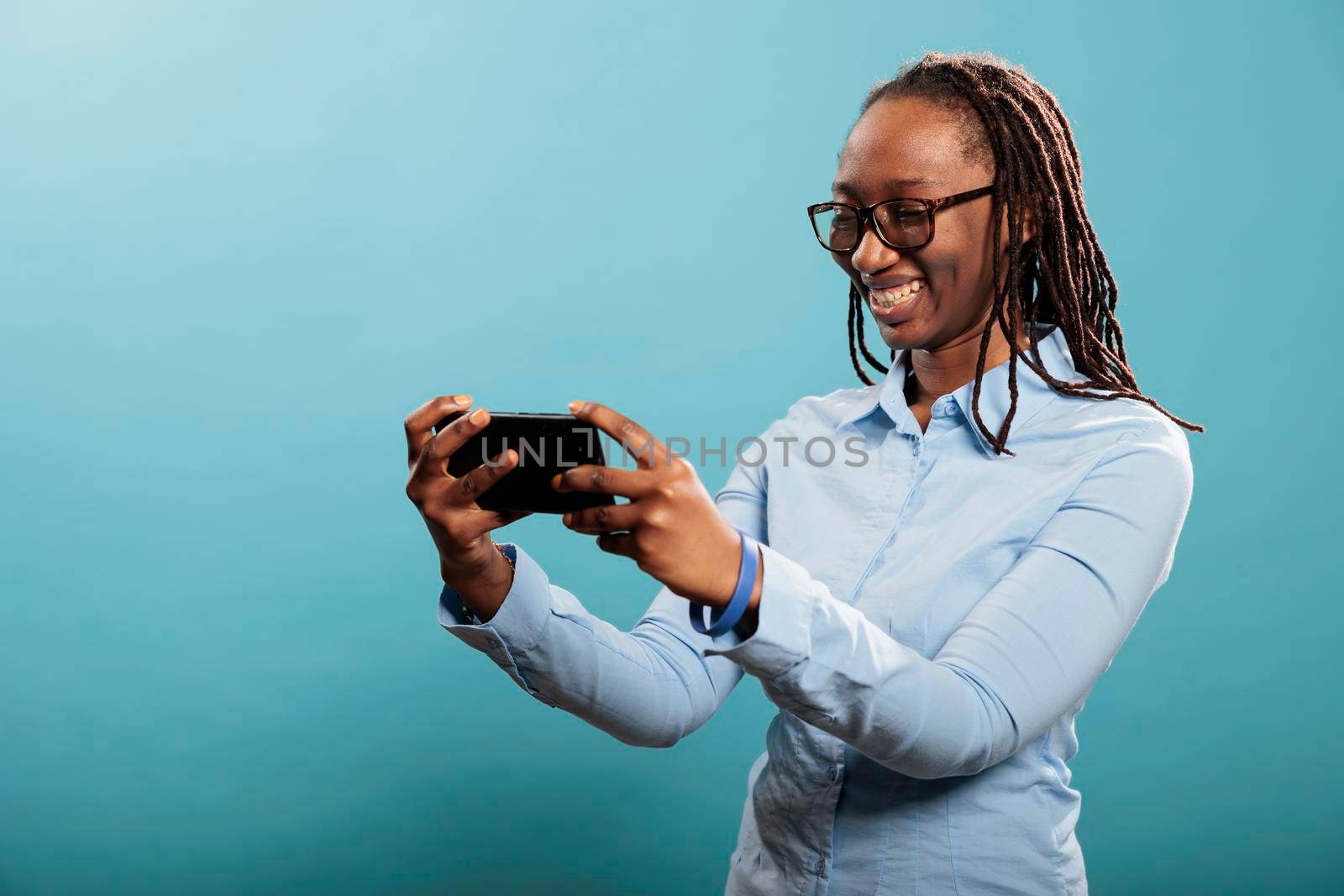 Confident joyful young adult with mobile cellphone browsing webpage on blue background. Happy smiling heartily woman modern smartphone touchscreen device enjoying watching funny videos on internet