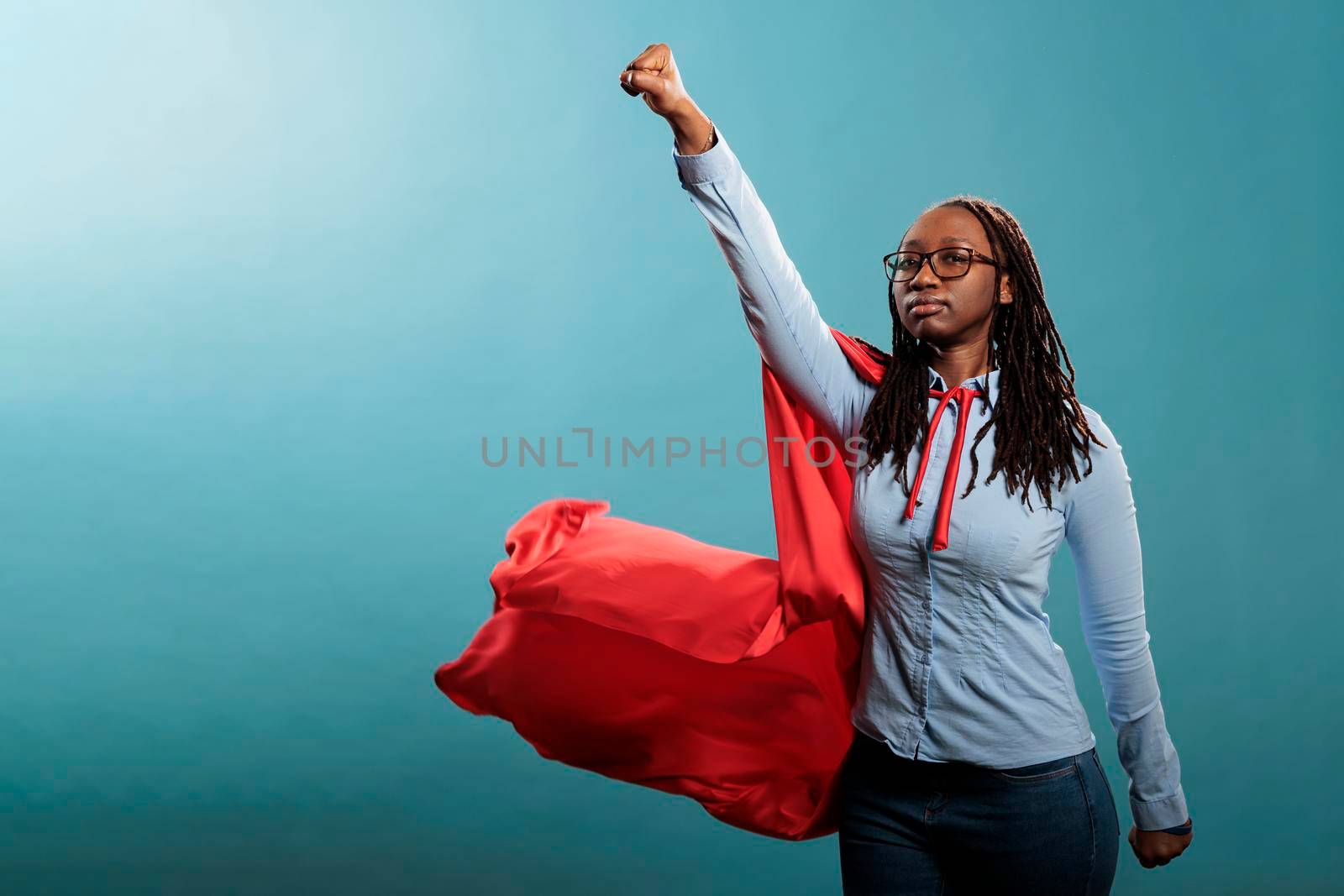 Mighty and brave african american superhero woman posing as justice defender while acting like flying on blue background. Young adult strong person wearing red hero cloak. Studio shot.