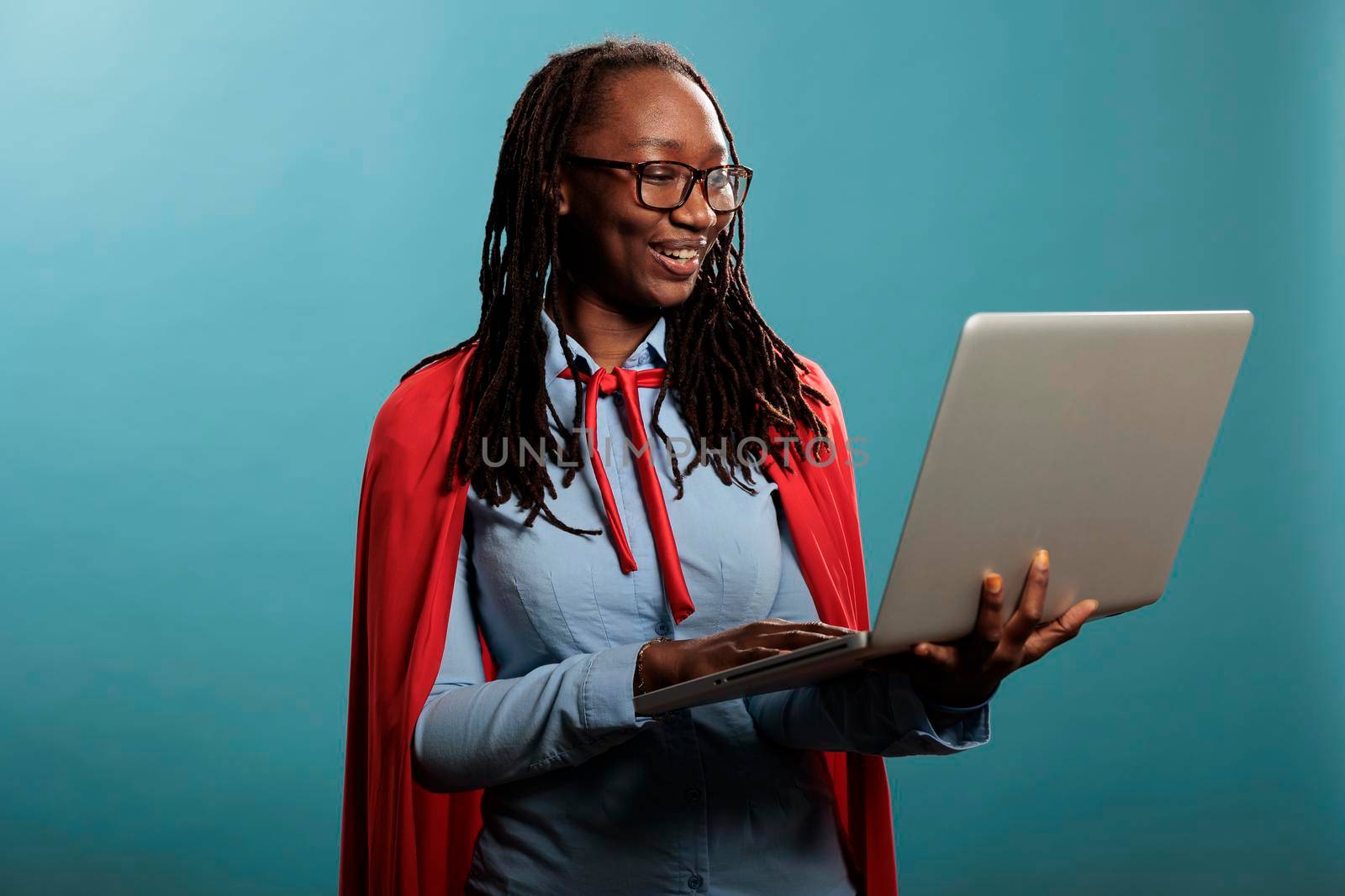 Confident and joyful justice defender wearing mighty hero cape while having handheld computer. Brave and proud young adult superhero woman using modern laptop while standing on blue background.