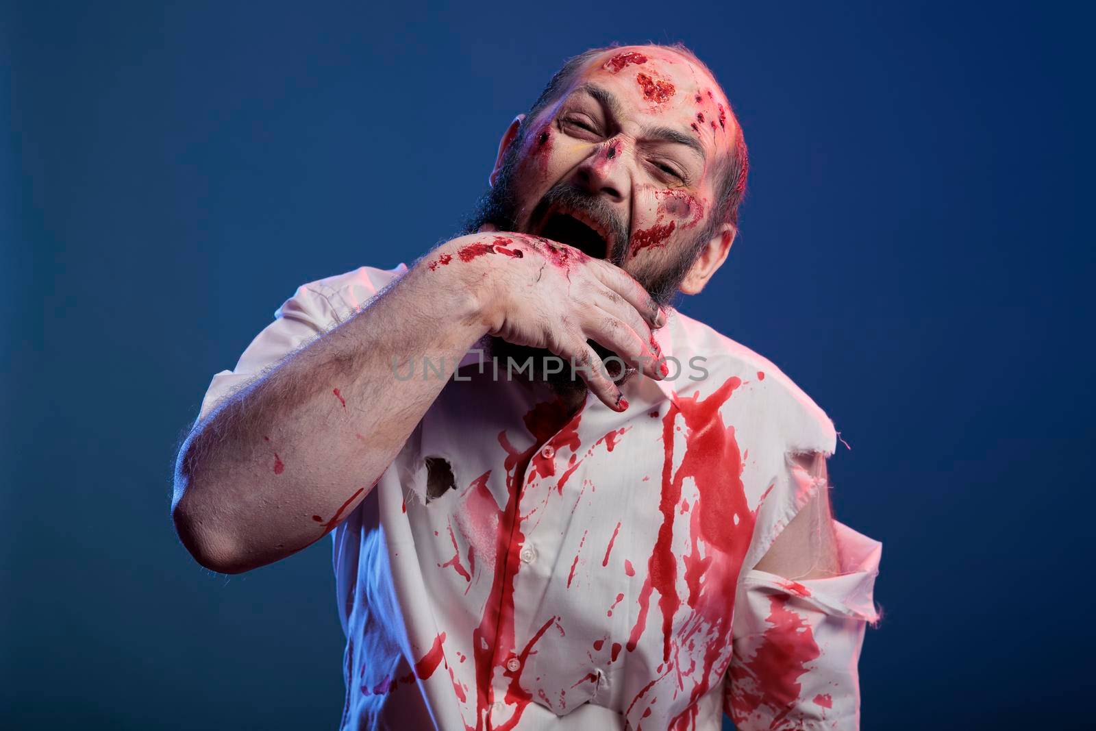 Halloween cruel zombie biting hand with scars and bloody wounds, standing in studio. Apocalyptic frightening corpse and brain eating villain with deadly horror face and aggressive sinister eyes.