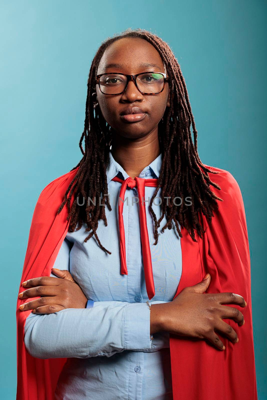 Mighty justice defender woman wearing superhero red cape standing with arms crossed while looking at camera. Young adult hero looking tough on blue background. Studio shot.