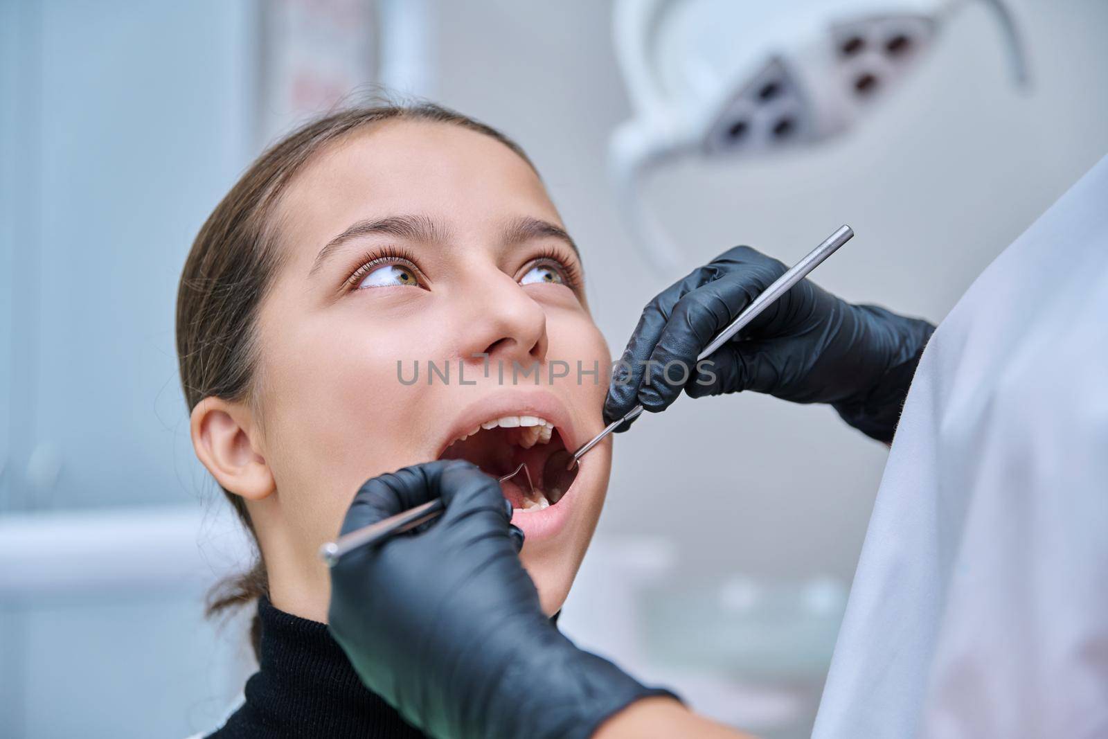 Young teenage female at dental checkup in clinic. Teenage girl sitting in chair, doctor dentist with tools examining patient's teeth. Adolescence, hygiene, treatment, dental health care