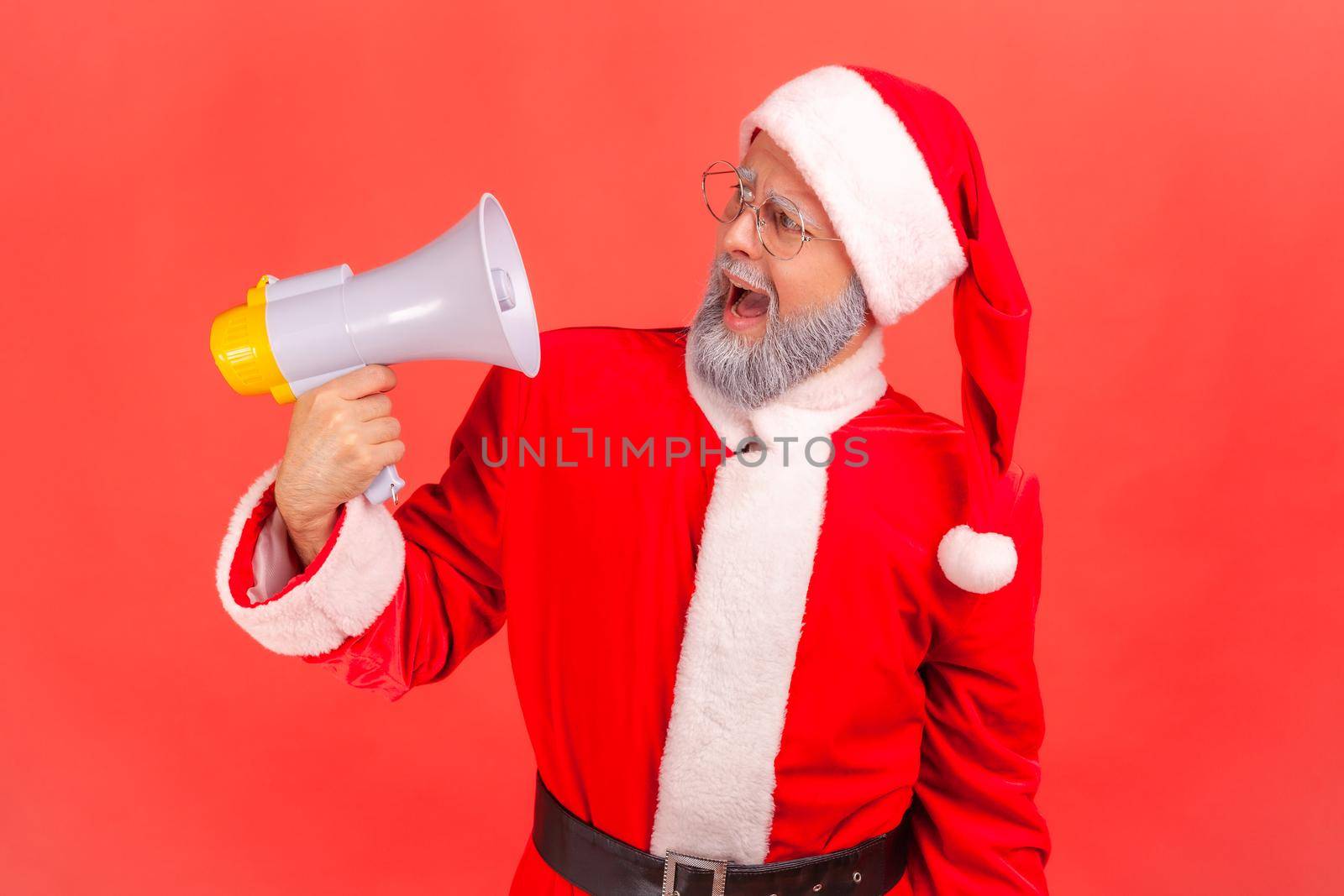 Portrait of crazy elderly man with gray beard wearing santa claus costume shouting loud, having angry facial expression, holding megaphone. Indoor studio shot isolated on red background.
