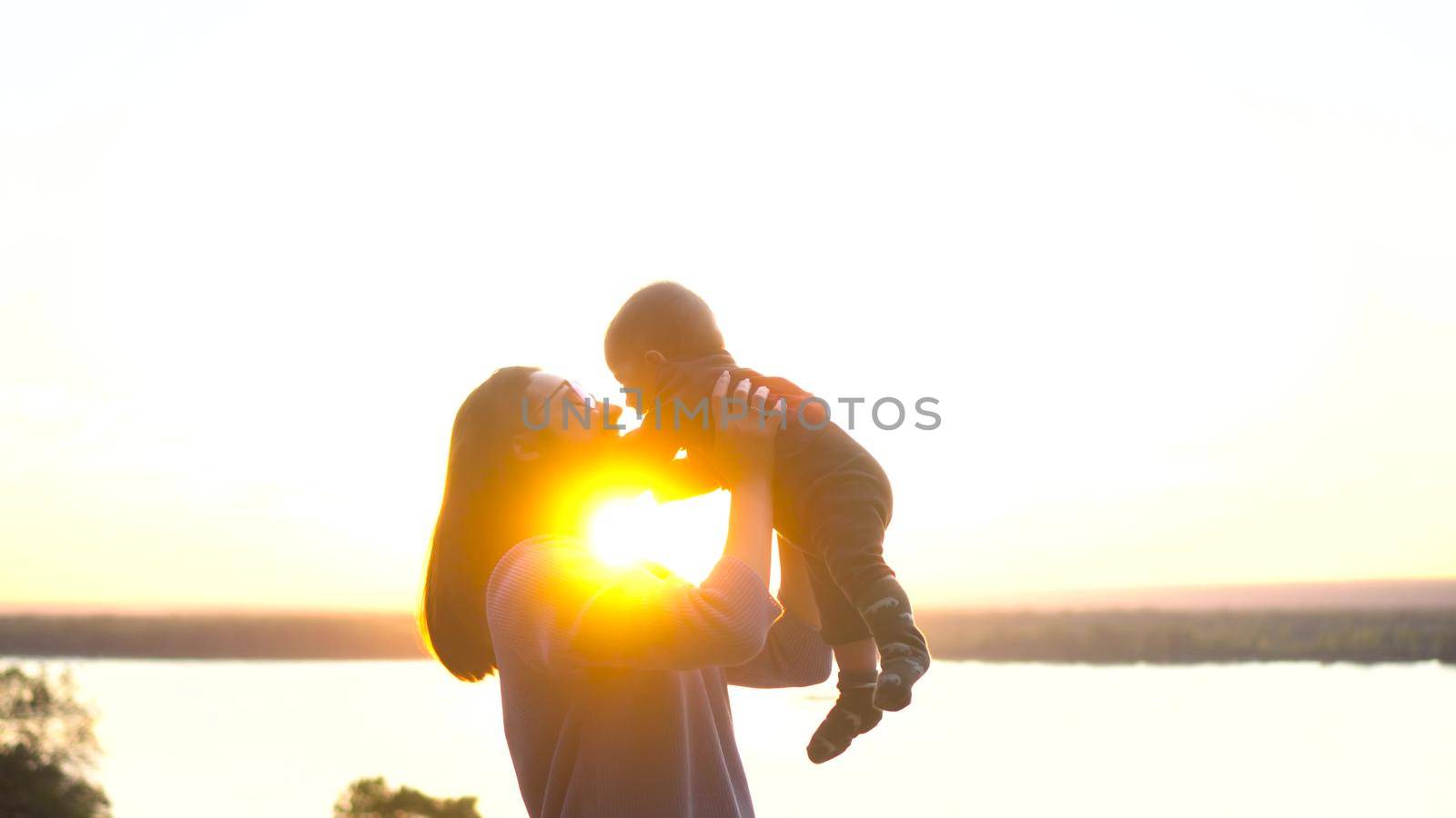 A young mother holds her baby son in her arms at sunset. A woman with glasses on a hill raises a child up against the background of the river. Close-up. 4k