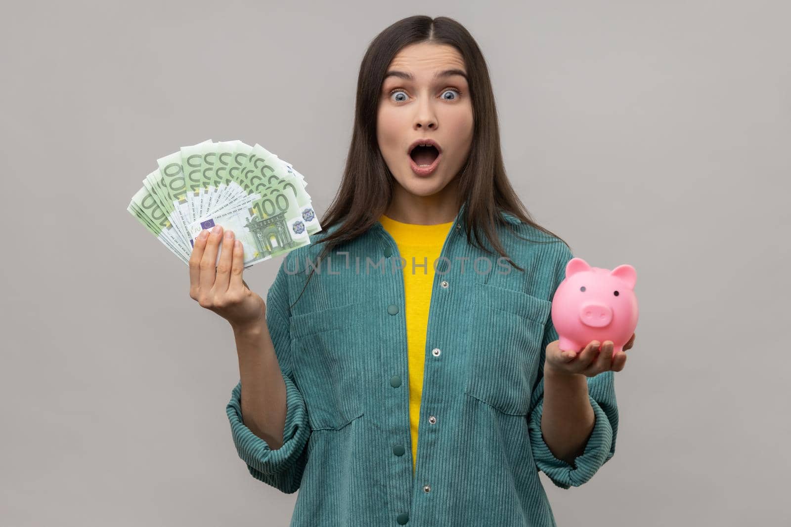 Portrait of shocked amazed woman holding big sum of euro banknotes and piggy bank, profitable investment, wearing casual style jacket. Indoor studio shot isolated on gray background.