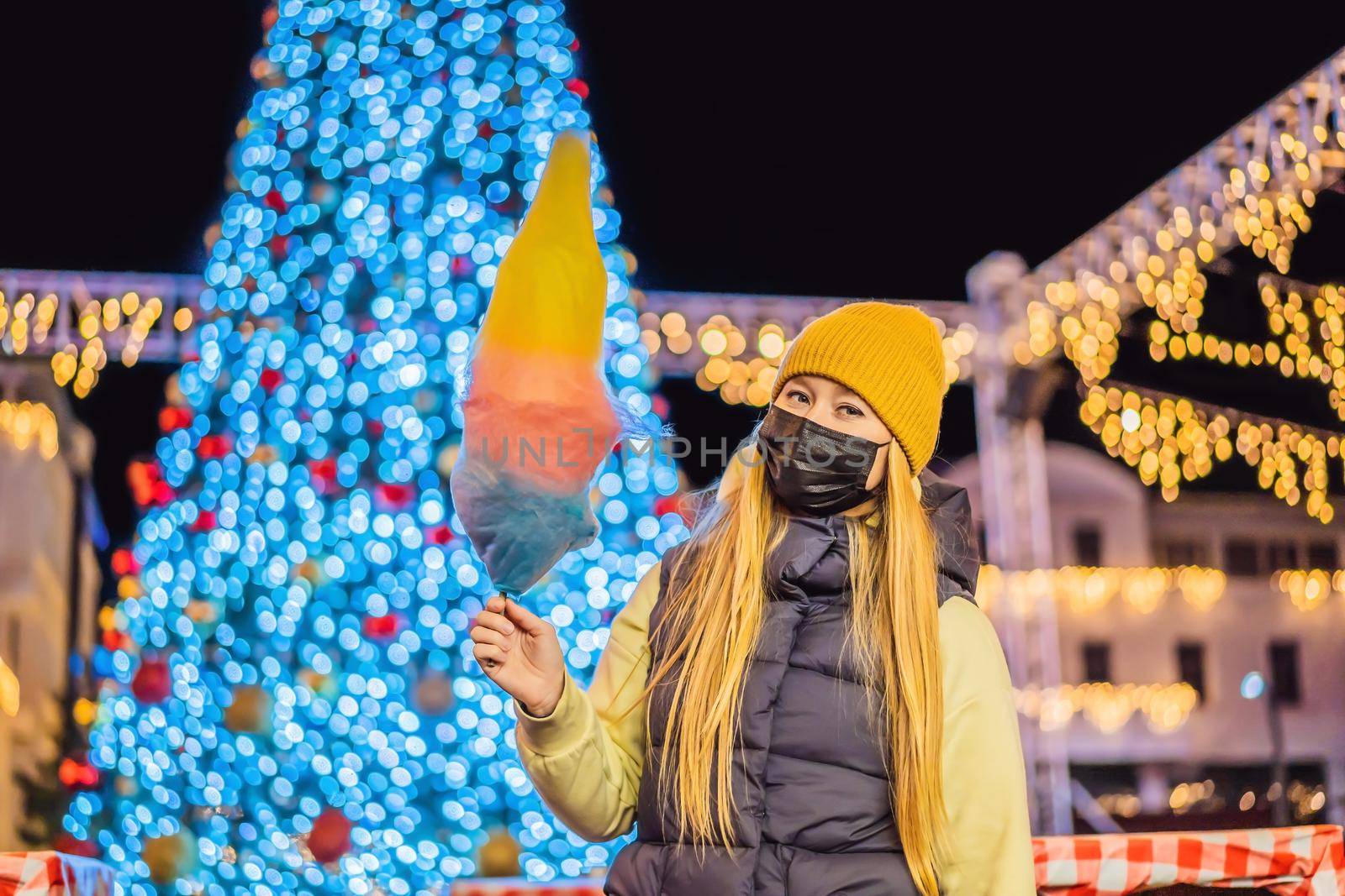 Young woman holding a cotton candy and smiling at a Christmas fair wearing a yellow wool cap wears a medical mask against coronavirus COVID 19 by galitskaya