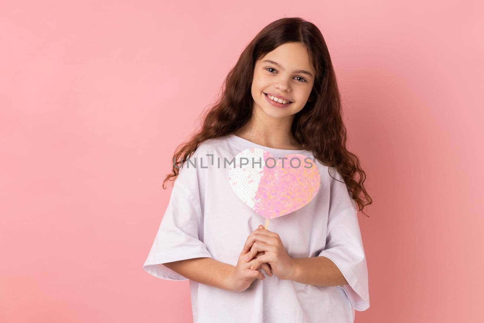 Portrait of happy satisfied little girl wearing white T-shirt holding paper pink heart on stick and smiling to camera, sharing love. Indoor studio shot isolated on pink background.