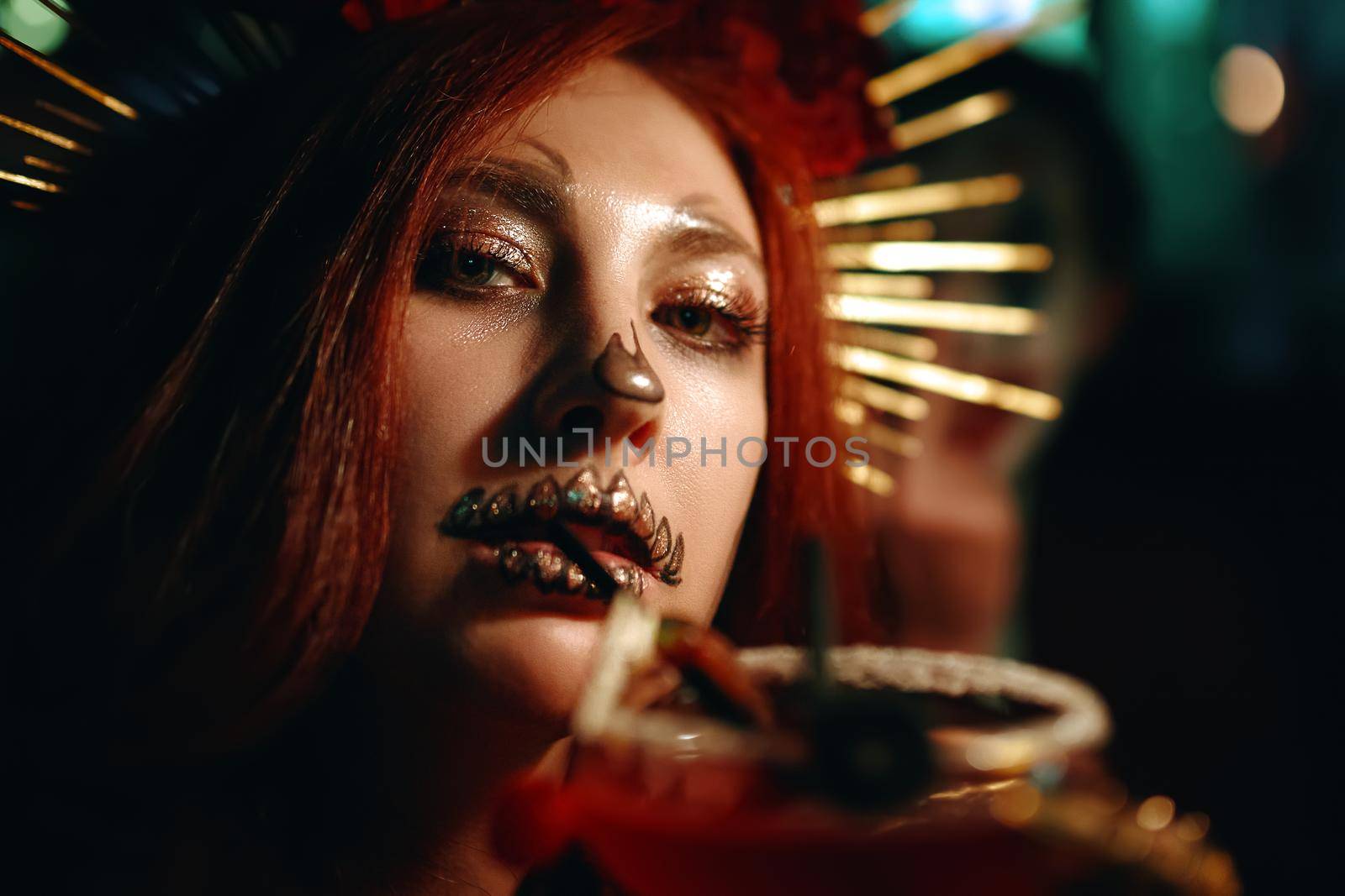 Woman with skeleton makeup at a Halloween party drinks cocktail and looks at camera. All Saints' Night. Celebration in a nightclub. Female in creepy costume of Mexican goddess of death.