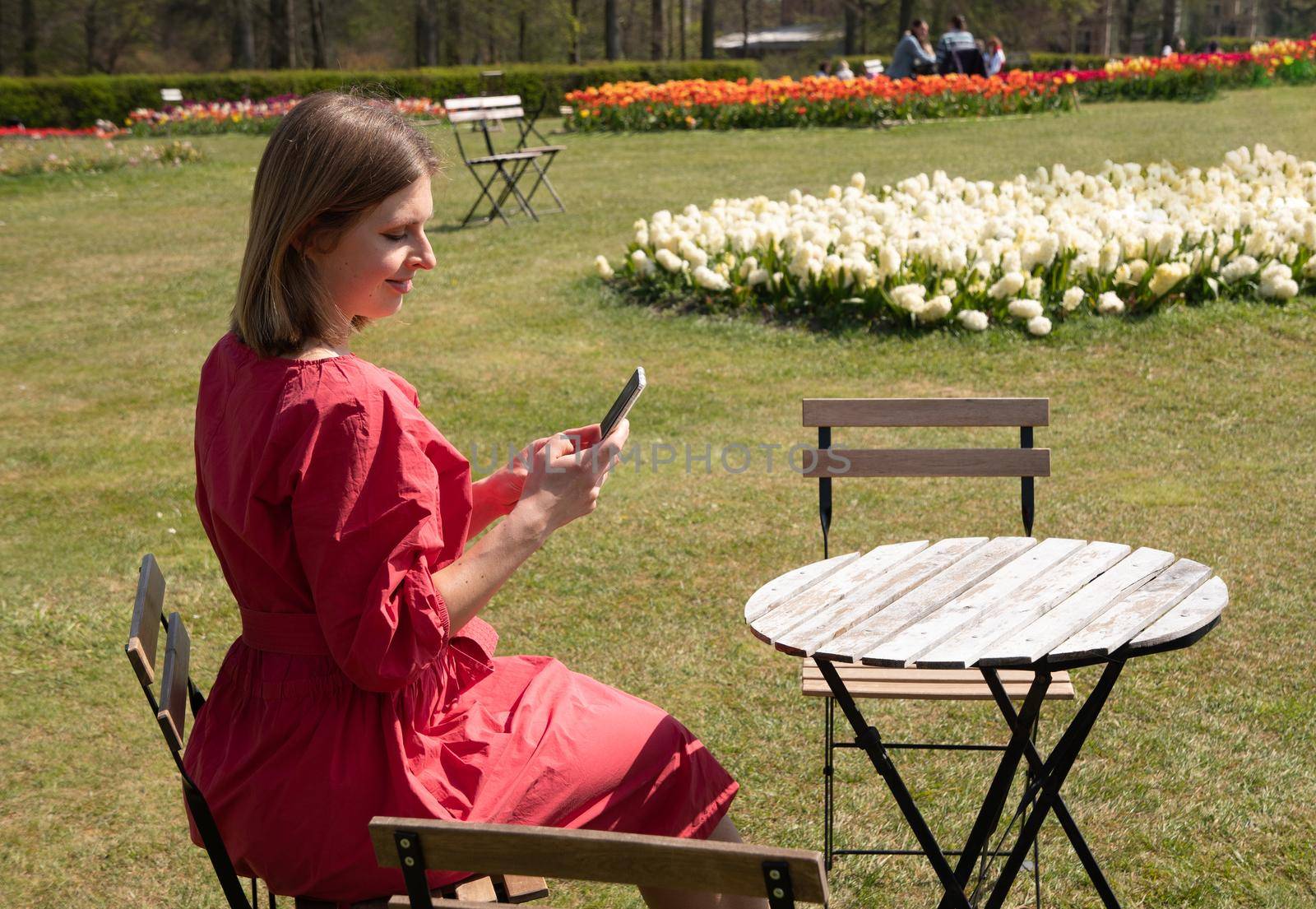 A young beautiful girl in a red dress in a spring park plays with her hair, flirts and takes a selfie on a mobile phone, tulips bloom and people walk in the park, pamper themselves.High quality photo