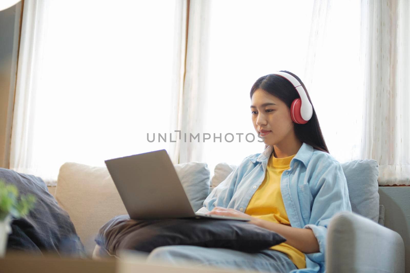 Young asian woman studying, online learing, and listening to music with headset while sitting on couch at home. Listening to podcast, audio book, watching videos using laptop.