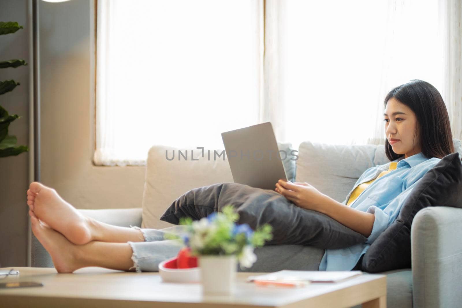 Young asian woman smiling and using laptop to study or work while leaning back on couch at home. Women online learning and searching for information using laptop while sitting comfortably on couch in the bright living room. Working, studying on desk at home. online learning