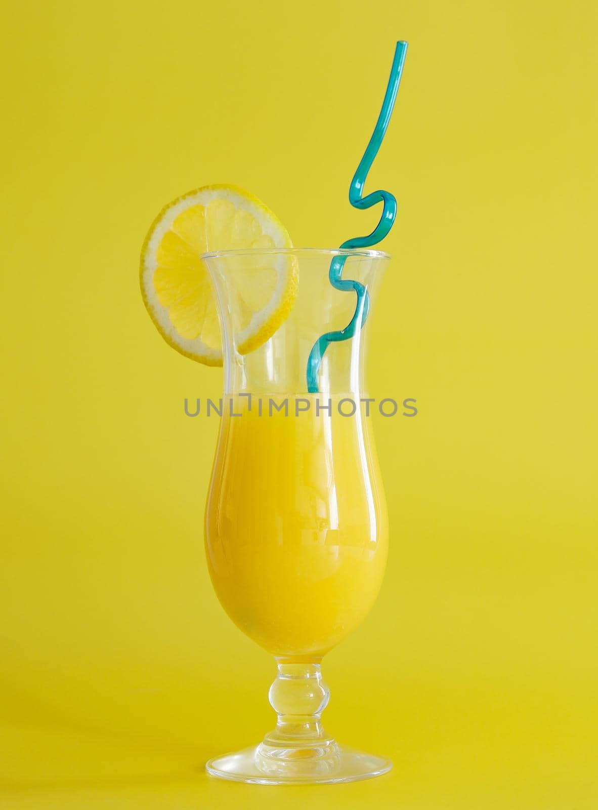 Fresh orange juice in glass, decorated with slice of lemon and blue straw. Summer cocktail on the yellow background. Colorful design