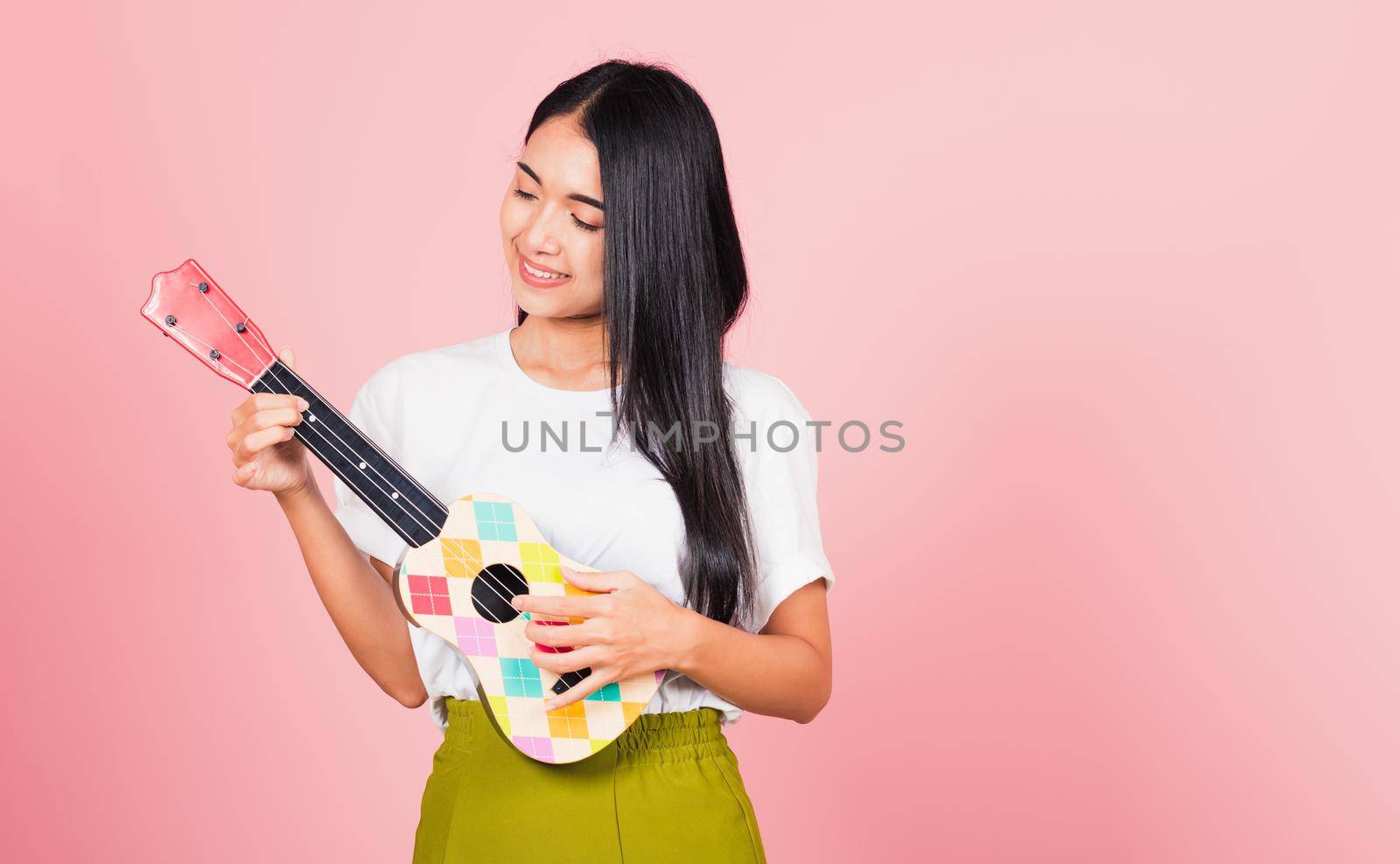 Portrait of happy Asian beautiful young woman teen confident smiling face hold acoustic Ukulele guitar, female playing Hawaiian small guitar, studio shot isolated on pink background, with copy space