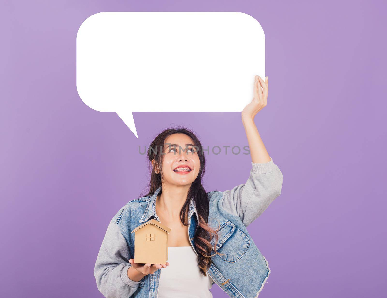 Happy Asian portrait beautiful cute young woman wear denim excited smiling hold house model and empty speech bubble sign, studio shot isolated on purple background, broker female real estate insurance