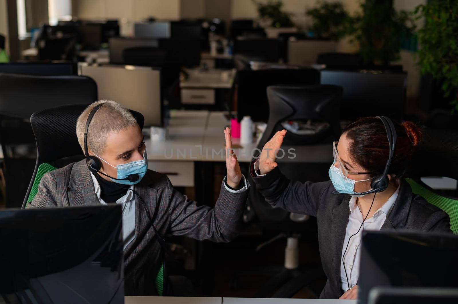 Two business women in masks are giving a high five while sitting at one desk in the office by mrwed54
