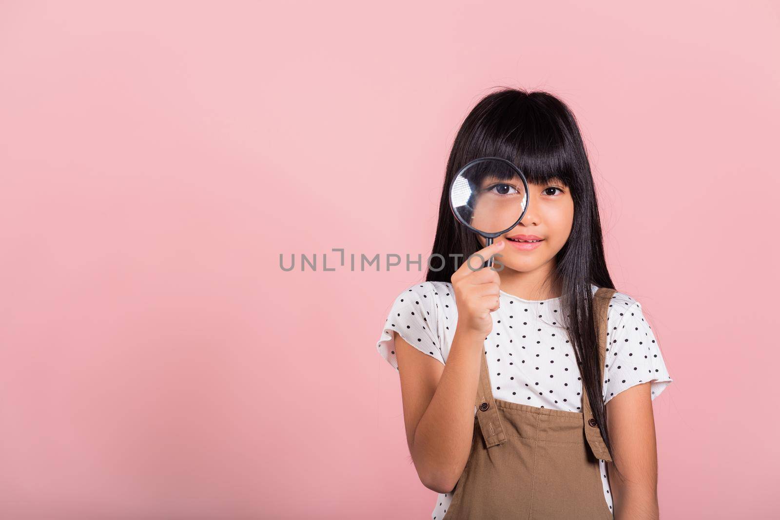 Asian little kid 10 years old funny looking through magnifying glass at studio shot isolated on pink background, Happy child girl lifestyle smiling exploring holding magnifier searching