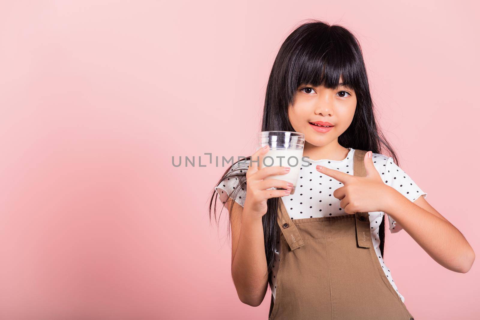 Asian little kid 10 years old smile hold milk glass drink white milk and pointing finger at studio shot isolated on pink background, Happy child girl daily life health care Medicine food