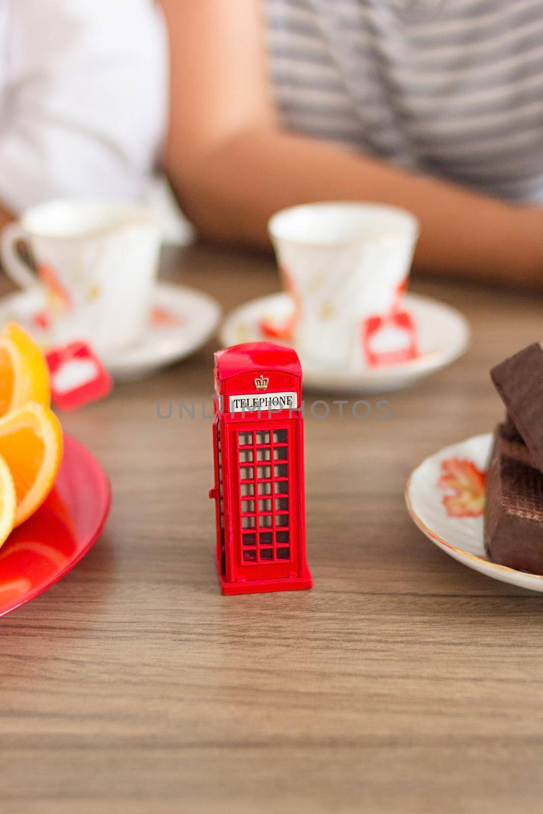 Traditional afternoon tea of british ceremony with such symbol of britishness as toy telephone box, defocused faceless women on background, front focus, vertical image