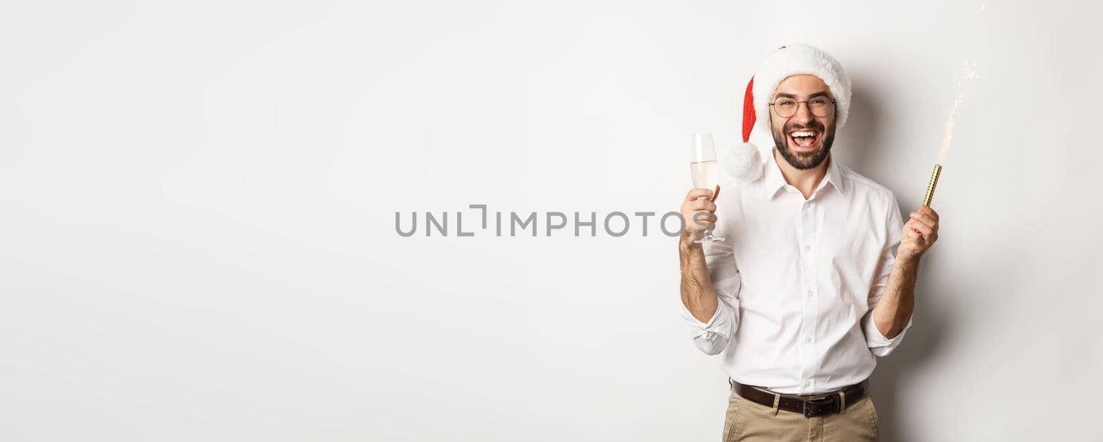 Winter holidays and celebration. Handsome bearded man having New Year party, holding firework sparkle and champagne, wearing santa hat, white background.