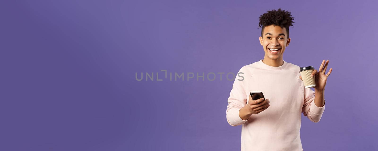 Portrait of cheerful young male student with cup of coffee and mobile phone, waving hand to say hi, smiling friendly as greeting person, meet classmate near university campus, purple background.