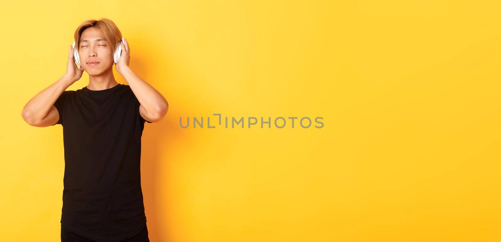 Relaxed and pleased handsome asian guy close eyes from satisfaction while listening music in wireless headphones, standing yellow background by Benzoix