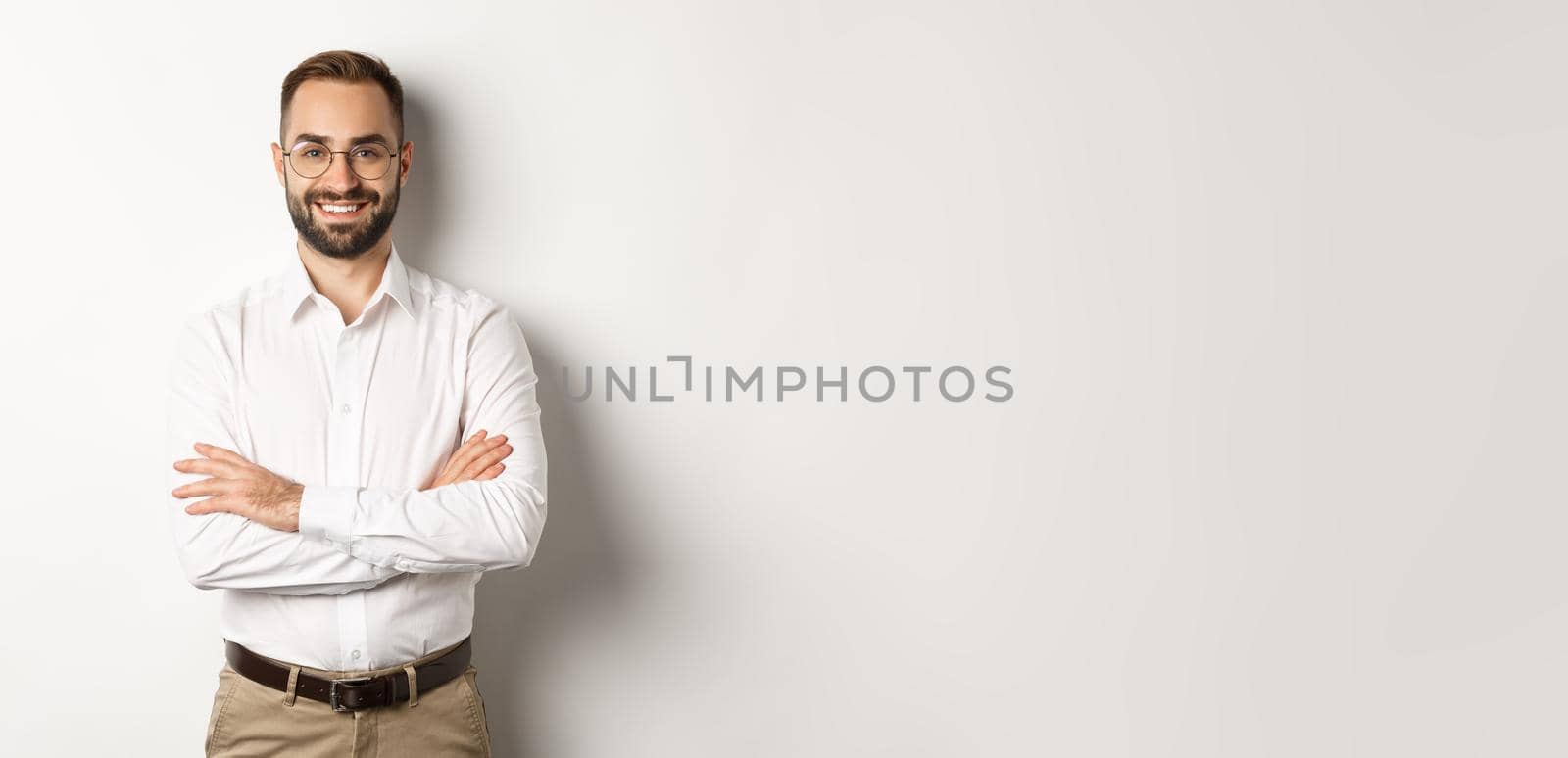 Business. Young professional businessman in glasses smiling at camera, cross arm on chest with confidence, standing over white background.