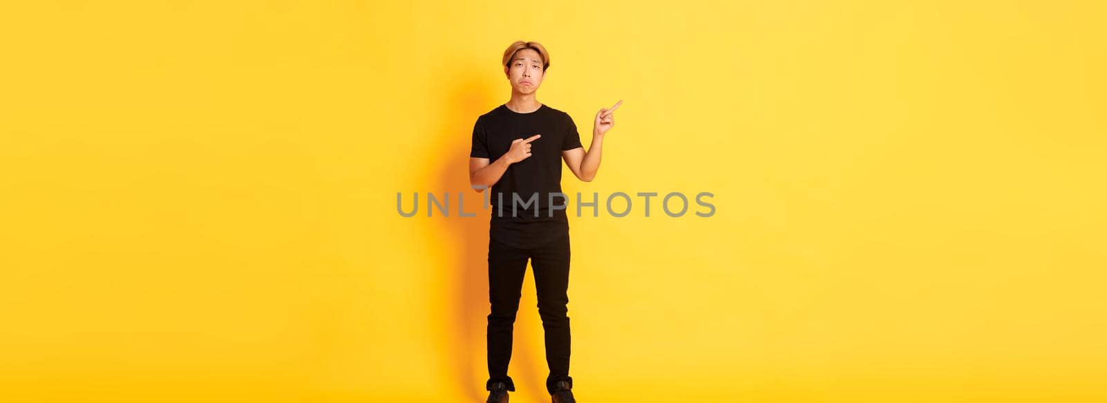 Full-length of sad handsome korean guy with blond hair making upset grimace, pointing fingers upper left corner, showing logo with regret, standing yellow background.