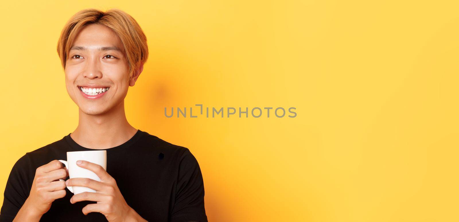 Close-up of happy smiling handsome blond asian guy, looking away with dreamy nostalgic look while drinking coffee, standing over yellow background.