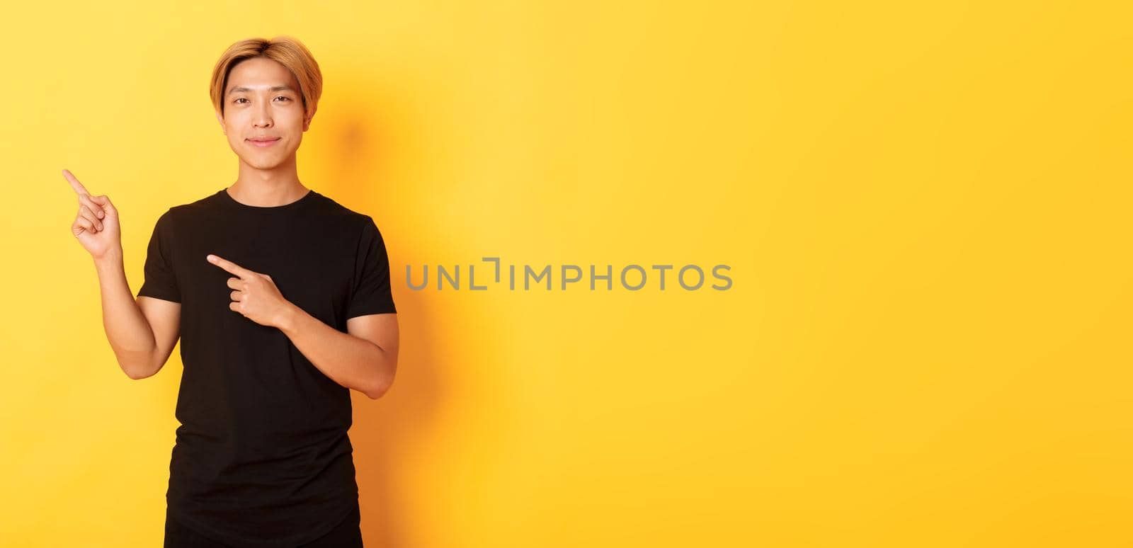 Portrait of smiling handsome asian man in black t-shirt pointing fingers upper left corner, showing logo or banner, yellow background by Benzoix