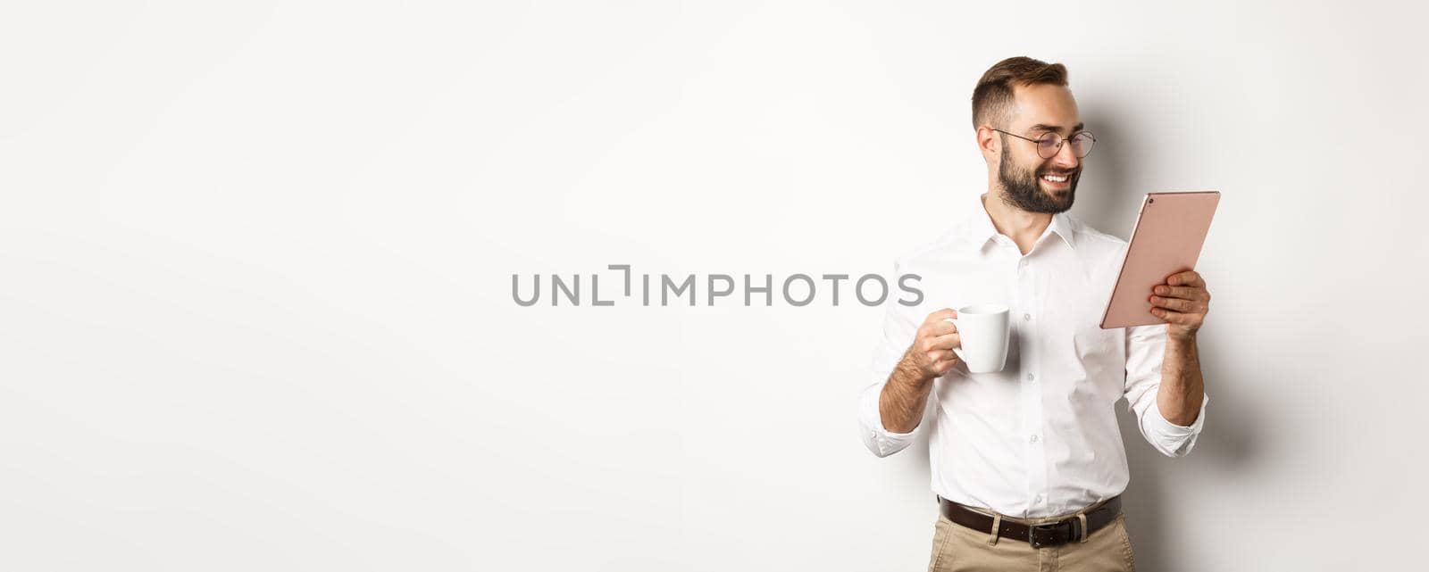 Handsome businessman drinking coffee and reading on digital tablet, smiling pleased, standing over white background.