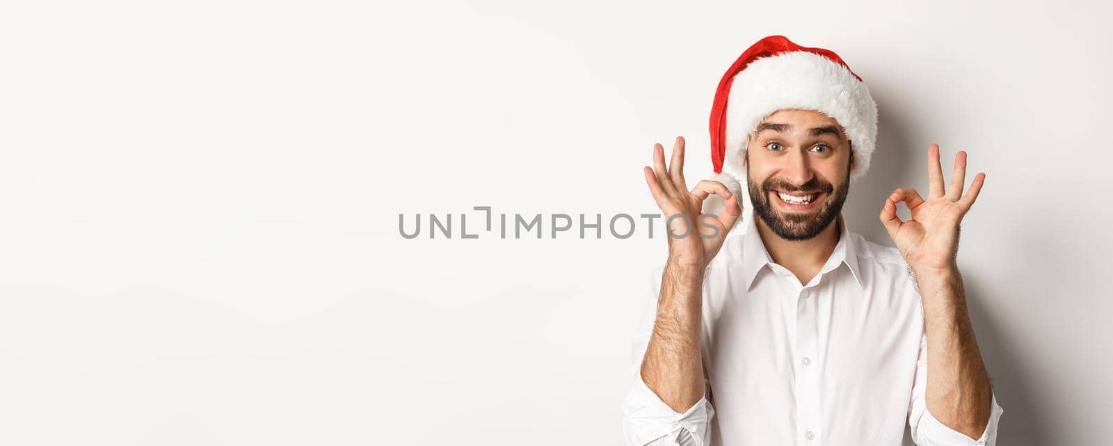 Party, winter holidays and celebration concept. Joyful man enjoying christmas and showing okay sign, smiling satisfied, wearing santa hat, white background.