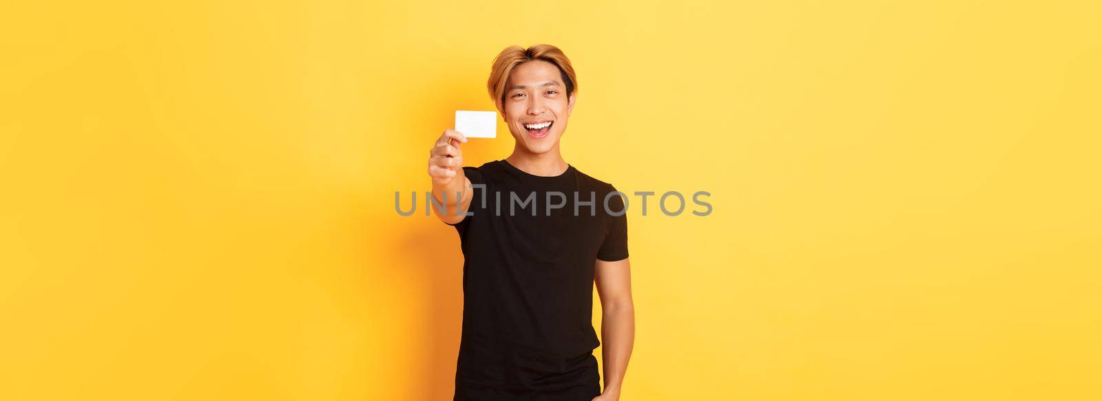 Cheerful handsome asian guy smiling pleased and showing credit card, standing yellow background by Benzoix