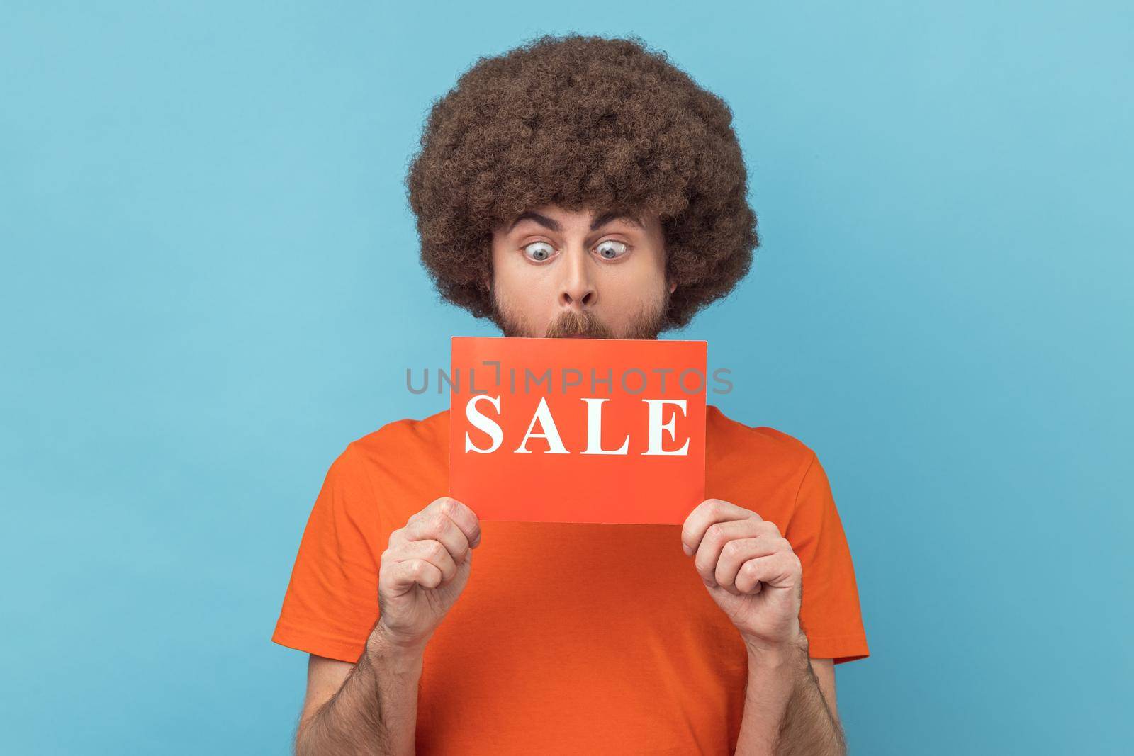 Man with Afro hairstyle wearing orange T-shirt standing with Sale word, looking with big eyes surprised, shocked by discounts, Black Friday low prices. Indoor studio shot isolated on blue background.