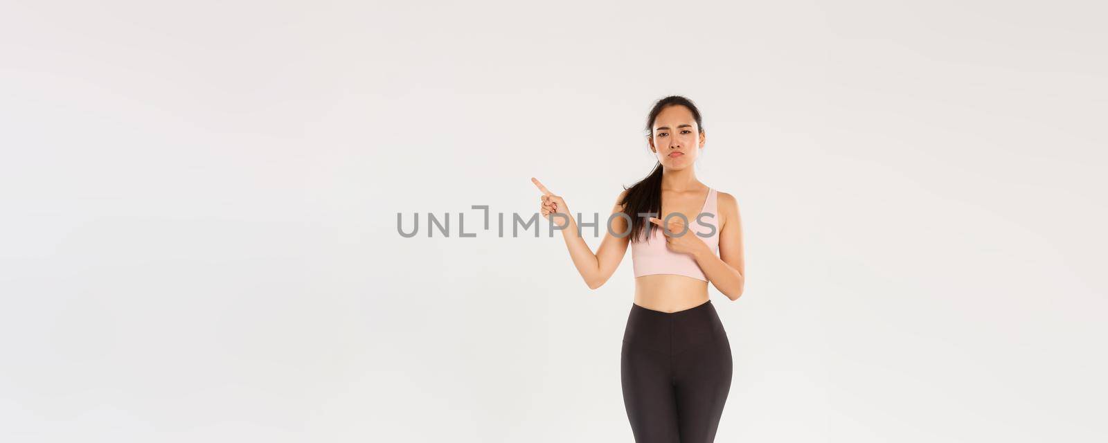 Full length of skeptical, disappointed asian fitness girl, sportswoman smirk and sulking while complaining on bad gym or awful workout gear, pointing upper right corner displeased, white background.