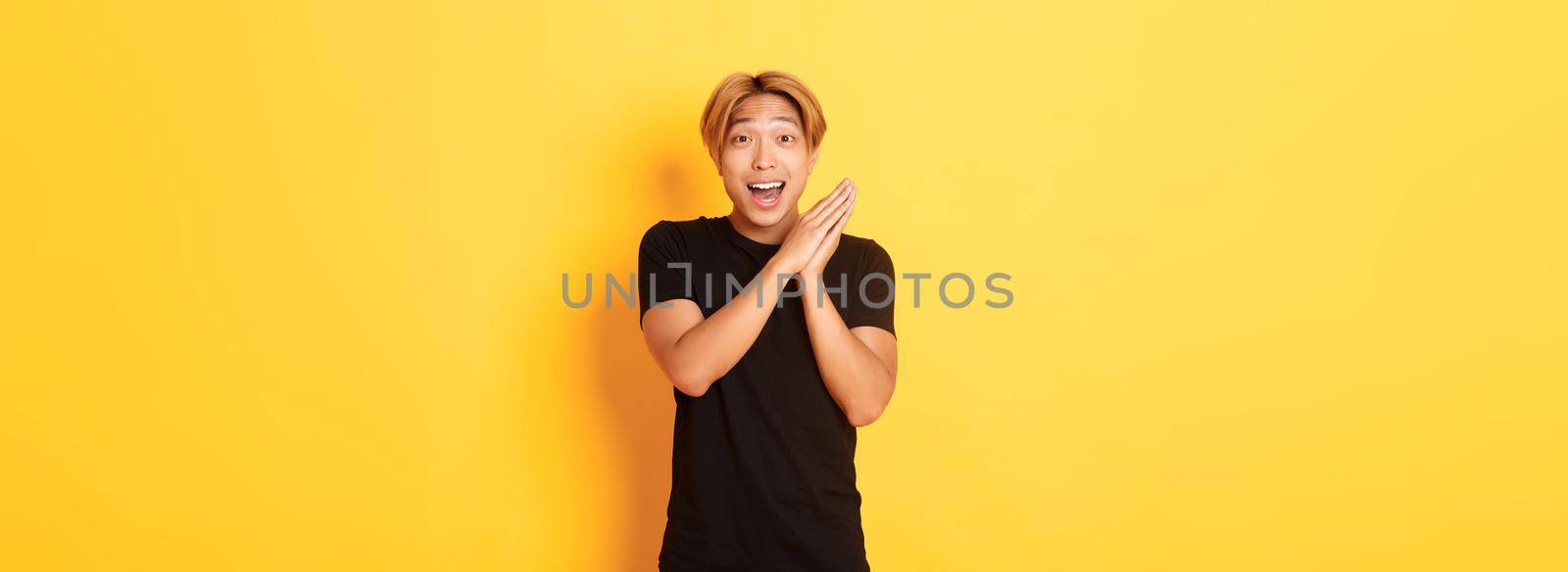 Portrait of happy and surprised asian man clap hands and smiling delighted, standing yellow background by Benzoix