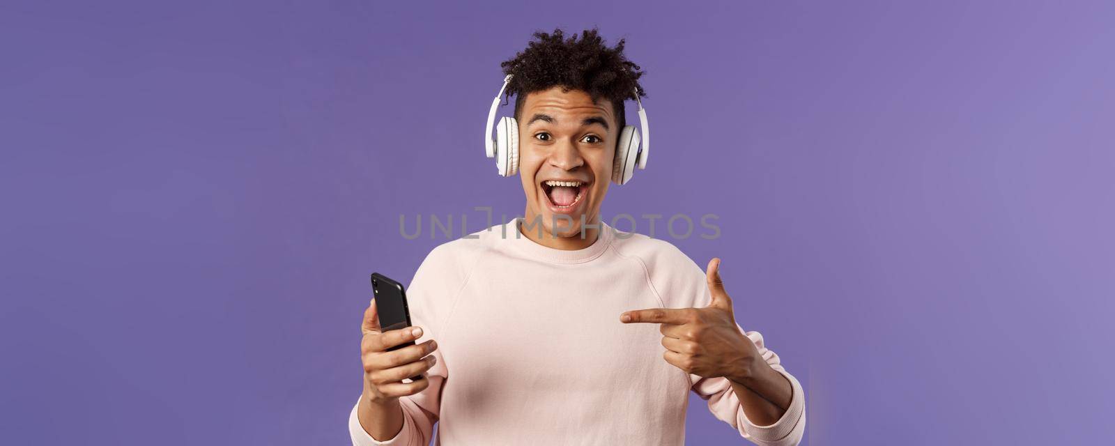 Technology and lifestyle concept. Portrait of happy, cheerful young man recommend awesome podcast or online music platform, buying subscribtion listen songs anytime, wear headphones, point at phone.