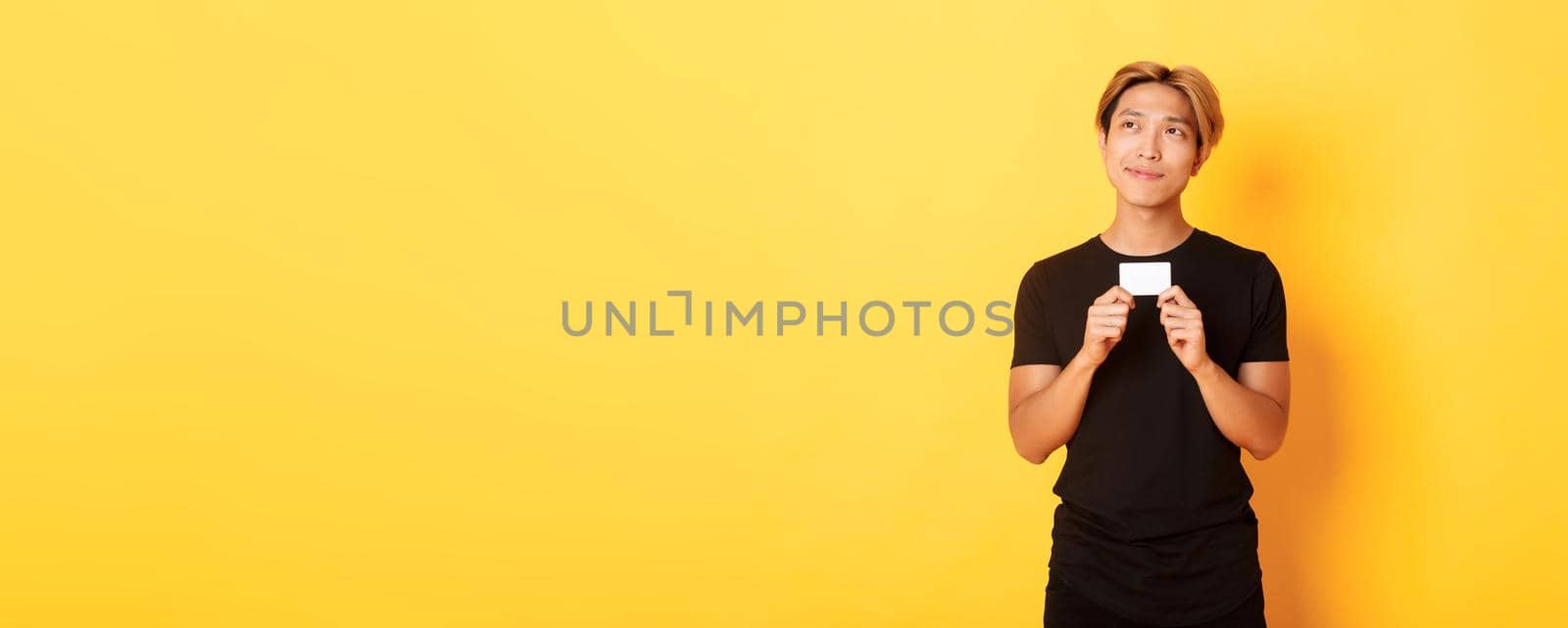 Thoughtful smiling asian guy thinking while showing credit card, looking upper left corner dreamy, yellow background by Benzoix