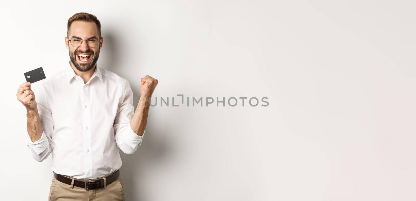 Satisfied handsome man showing credit card and rejoicing, winning something or triumphing, standing over white background.