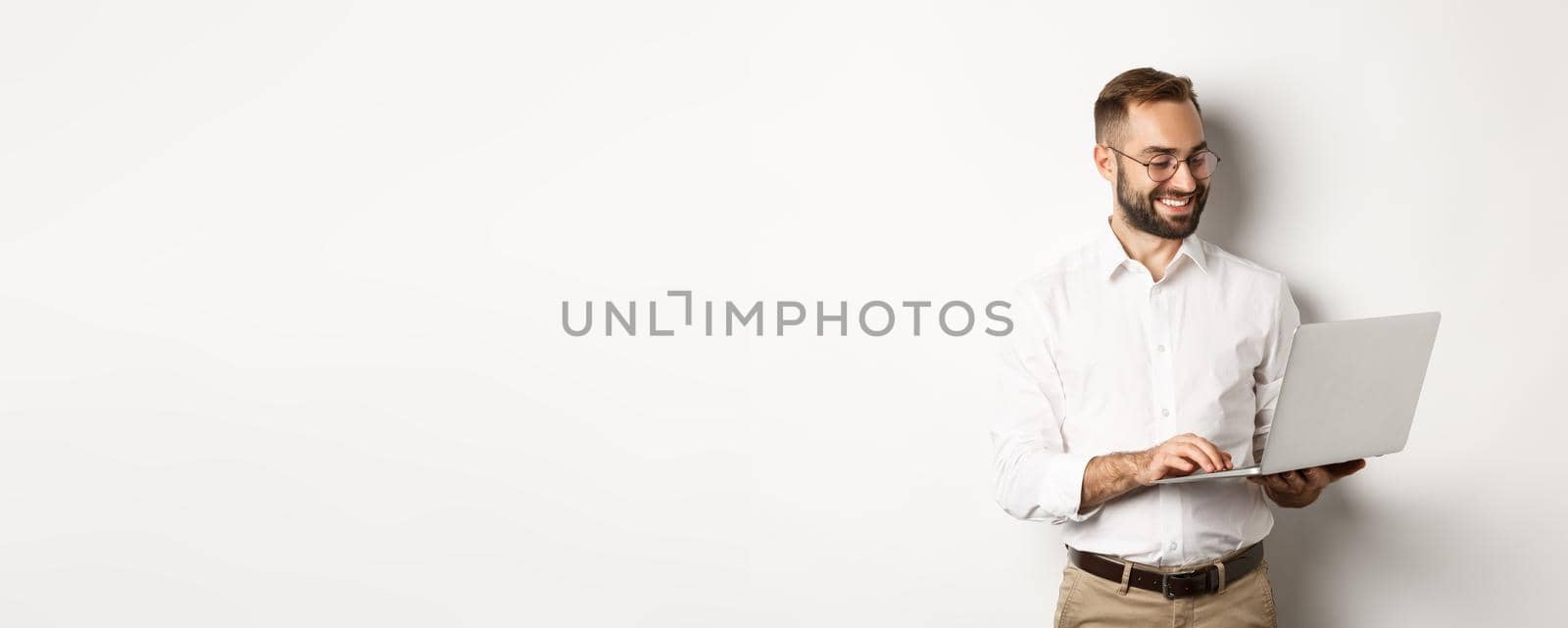 Business. Handsome businessman working on laptop, answering messages and smiling, standing over white background.