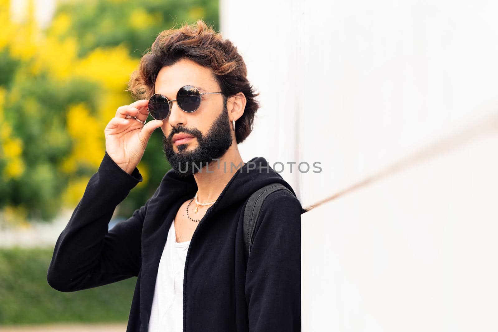 portrait of a beautiful young man putting on sunglasses leaning in a white wall, concept of urban lifestyle and stylish clothing, copy space for text