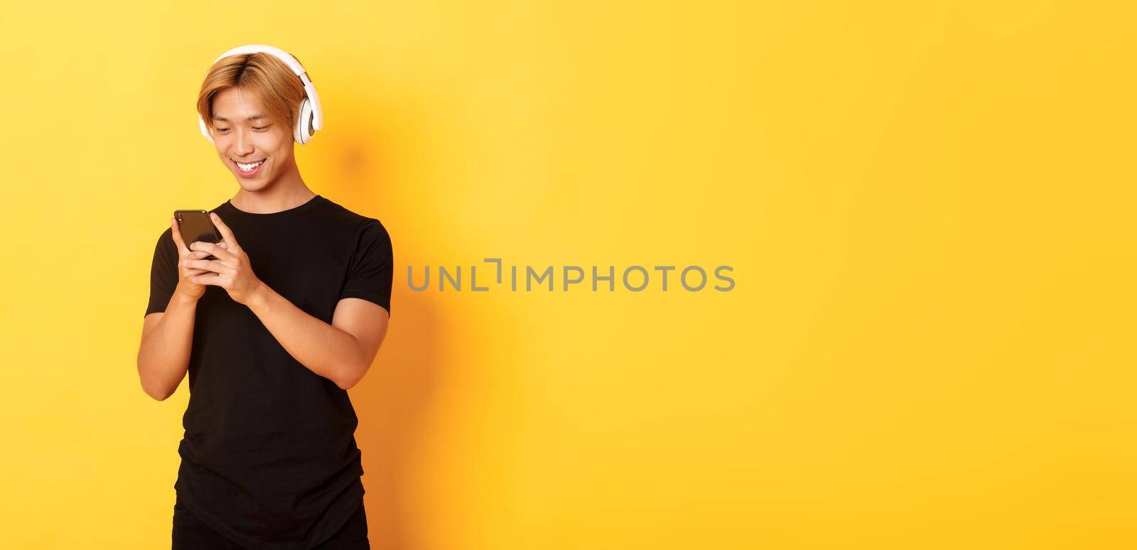 Young smiling asian guy using smartphone and listening music or podcast in wireless headphones, standing yellow background by Benzoix