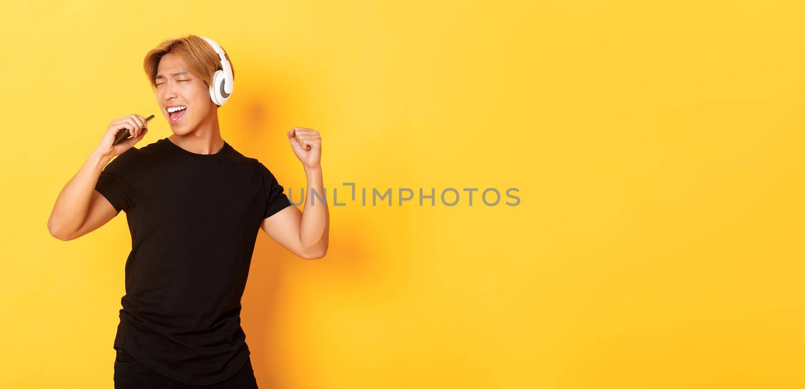 Carefree handsome asian guy in headphones, playing karaoke app, singing into mobile phone microphone, standing yellow background by Benzoix